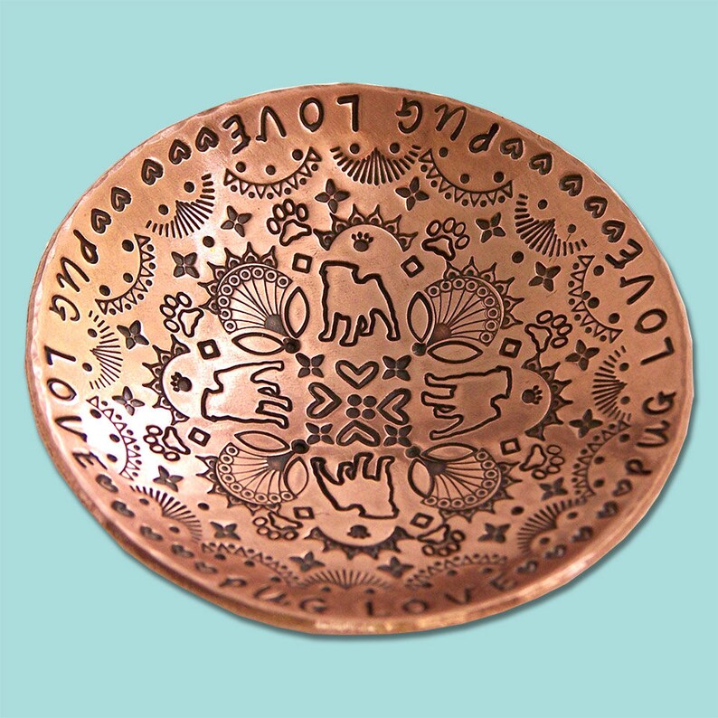 May include: A copper bowl with an intricate design featuring paw prints, hearts, and the words "Love Pug Love" repeated around the rim.
