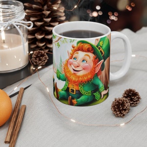 May include: A white ceramic mug with a handle features a colorful illustration of a smiling leprechaun wearing a green hat and holding a pot of gold. The mug is filled with a dark liquid.