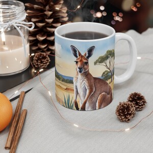 May include: A white ceramic mug with a brown kangaroo illustration. The kangaroo is standing in a field with green grass and a blue sky in the background. The mug is filled with a dark brown liquid.
