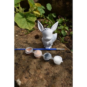 May include: A white unpainted Eevee figurine, a blue-handled paintbrush, and a strip of four small plastic paint pots containing brown, peach, black, and white paint. The figurine features pointed ears and a fluffy collar, presented as a craft kit.