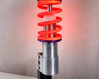 Op de afbeelding: Industriële lamp met een rode spiraalveer, zilverkleurige metalen basis en zwarte cirkelvormige standaard. De lamp geeft een rode gloed, met paarse en oranje accenten. Modern ontwerp, ongeveer 30 cm hoog.
