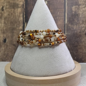 May include: A multi-strand beaded bracelet in shades of brown, gold, and cream, displayed on a white cone-shaped stand with a wooden base. The beads vary in size and texture, creating a layered effect. The background is a wooden surface.