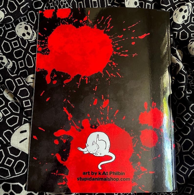 May include: A black book cover with large red splatters and a white cat illustration giving the middle finger. The text "art by kAt Philbin stupidanimalshop.com" is printed at the bottom.