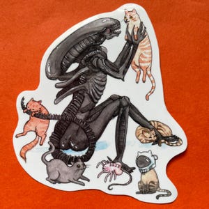 May include: A sticker featuring a black Xenomorph from the Alien film franchise interacting with several cats. The cats are various colors, including orange tabby, gray, and a cat wearing a space helmet. The sticker is on an orange background.