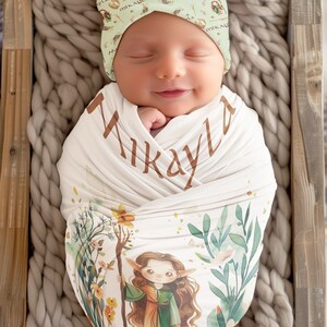 May include: A white swaddle blanket featuring a green and brown illustration of a fairy holding a staff. The blanket is wrapped around a baby wearing a green and white hat with the name "Mikayla" printed on it.