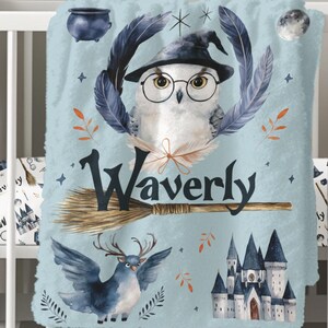 May include: A blue baby blanket with a white owl wearing a witch hat and glasses, holding a broom with the name "Waverly" written on it. The blanket also features a blue bird with antlers, a castle, and other magical elements.