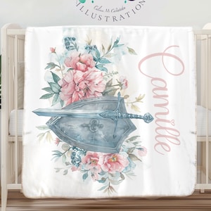 May include: A white baby blanket featuring a watercolour design of pink and blue flowers, a sword, and a shield. The name "Camille" is written in pink script on the right side of the blanket.