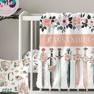 May include: A baby bedding set featuring a white blanket with the name "Cassandra" in a peach band, and swords decorated with pink roses. A matching pillow and crib bumper with floral and castle designs complete the set.