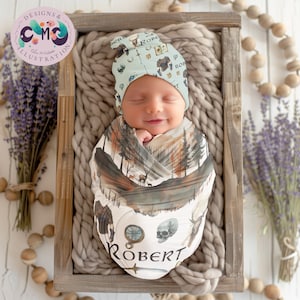May include: A newborn baby swaddled in a white and brown swaddle with a forest scene and the name "ROBERT" printed on it. The baby is wearing a light blue and green hat with a woodland creature and sword design.
