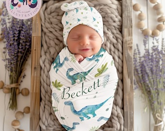 May include: A white swaddle blanket with a green dinosaur print and the name "Beckett" written in green. A baby is wrapped in the blanket and wearing a matching green dinosaur print hat.