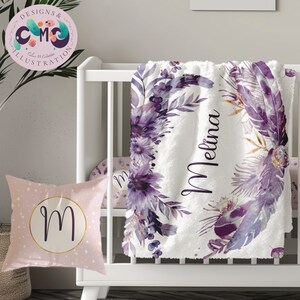 May include: A baby blanket, pillow, and crib sheet set. The white blanket features a floral and feather design in shades of purple, with the name "Melina" in script. The pillow is pink with a gold circle and the letter "M".