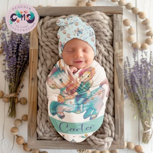 May include: A baby swaddled in a white and blue swaddle featuring a unicorn and a boy riding it. The swaddle is personalised with the name "Carter". The baby is wearing a blue and white hat with a star and unicorn pattern.