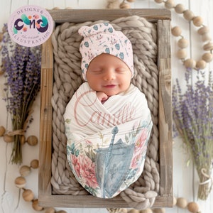 May include: A pink and white swaddle with a floral and sword design. The swaddle is wrapped around a sleeping baby wearing a pink and white hat with a similar design. The name "Camille" is written on the swaddle.