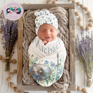 May include: A white swaddle blanket featuring a watercolour design of a sword, shield, and flowers. The blanket is wrapped around a sleeping baby wearing a white hat with a similar design. The baby's name, "Michael", is written on the blanket.
