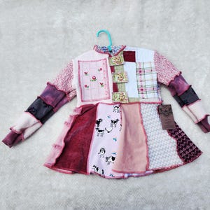 Könnte beinhalten: Ein Patchwork-Kleid mit verschiedenen Stoffen, darunter rosa Blumenmuster, Karomuster und Kuhmuster. Die Ärmel sind eine Mischung aus lila und rosa Farbtönen. Das Kleid hängt an einem blauen Kleiderbügel.