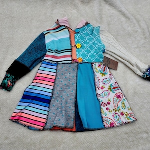 Könnte beinhalten: Ein Patchwork-Kleid mit einer Vielzahl von Stoffen in Türkis, Blau, Rosa und Weiß. Das Kleid hat lange Ärmel, einer in einem Zopfmuster und der andere in einem türkisfarbenen Strick. Das Oberteil ist mit orangefarbenen und gelben blütenförmigen Knöpfen verziert.