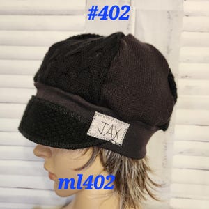 May include: Black knit beanie hat with a cable knit design on the crown and a textured brim. A small white tag with the name "JAX" is sewn on the brim. The text "#402" and "ml402" are in blue.