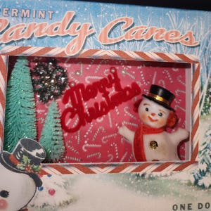 May include: A Christmas diorama with a snowman wearing a black top hat and red scarf, set against a pink backdrop with candy cane motifs. The scene includes miniature green Christmas trees and a red "Merry Christmas" sign, evoking a festive holiday atmosphere.