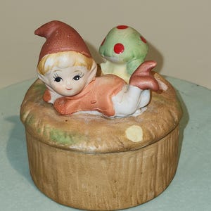 May include: A ceramic trinket box with a whimsical design. A small girl with a red hat and blonde hair is lying down next to a green mushroom with red spots. The box is decorated with a brown and green woodland scene.