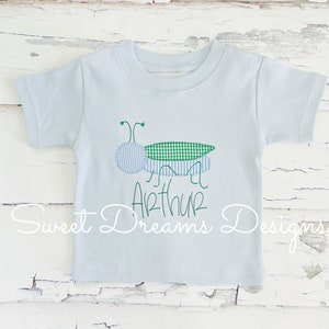 May include: A light blue t-shirt with an embroidered green and blue grasshopper and the name "Arthur" in green cursive writing. The shirt is from Sweet Dreams Designs.
