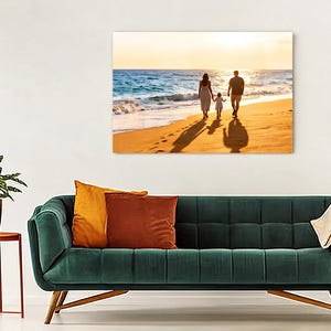 May include: A rectangular wall art print featuring a sunset beach scene with three figures walking along the shoreline. The print is displayed above a dark green tufted sofa with orange and rust-coloured throw pillows and a cream-coloured blanket.