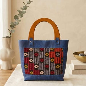 May include: A blue handbag featuring a central rectangular panel with a woven geometric pattern in red, gray, and yellow. The bag has a semi-circular wooden handle attached with blue leather tabs and silver-toned metal rivets at the top.