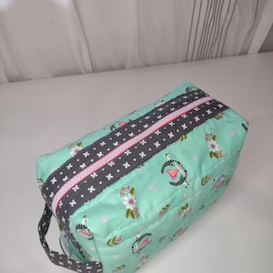 May include: A rectangular, quilted cosmetic bag in a light teal fabric with a floral pattern. A dark gray band with white cross accents and a pink zipper runs across the top. A small gray handle is attached to the side.