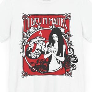 Puede incluir: Camiseta blanca con un estampado gráfico rojo, negro y blanco de una mujer sosteniendo una manzana con una serpiente enroscada a su alrededor. El texto "Ni Dieu Ni Maitre" está impreso en negro en la parte superior del gráfico.