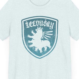 May include: A light blue t-shirt with a distressed blue and white graphic of a winged creature with a crown. The text "ŽELVIŠKÝ" is above the creature.