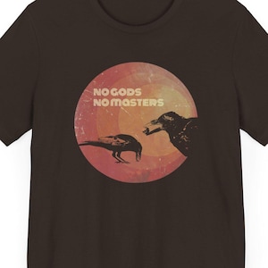 May include: A brown t-shirt with a circular design featuring two crows and the text "NO GODS NO MASTERS" in white.