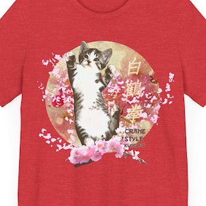 Puede incluir: Camiseta roja con un gato blanco y gris en una pose de grúa rodeado de flores de cerezo rosas. El texto "Crane Style Kung Fu" está impreso en la esquina inferior derecha de la camiseta. El texto "白鶴拳" está impreso en el centro de la camiseta.