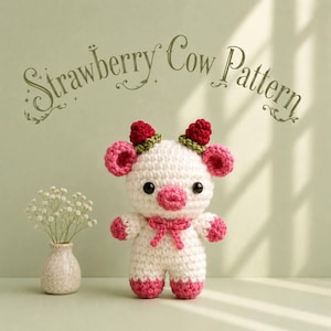 May include: A white and pink crocheted cow plushie with red strawberry-shaped horns, a pink snout, and a small pink bow at the neck. The text Strawberry Cow Pattern is displayed above the plushie, which stands next to a small vase of flowers.