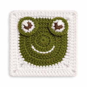 May include: A square, crocheted frog face with a white border. The frog's face is green with brown and white eyes and a smiling mouth. The crochet stitches are visible, giving it a handmade appearance.