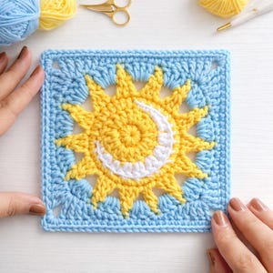May include: A square, crocheted sun and moon design in yellow, white, and blue. The sun is in the center, with a white crescent moon. The square is surrounded by a blue border. The yarn is visible in the background.