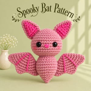 May include: A crocheted bat plush toy in shades of pink with black safety eyes, a small pink nose, and bright pink inner ears. The wings feature pink embroidered lines. The text Spooky Bat Pattern is displayed above the bat in dark green.
