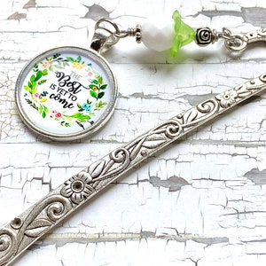 Peut inclure: Marque-page argenté avec des détails floraux et des volutes. Un pendentif rond avec le texte "The Best is Yet to Come" est attaché. Le pendentif est entouré d'une couronne florale avec des fleurs colorées. Un accent de perle et de fleur blanc et vert.