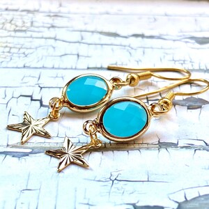May include: Gold-tone earrings featuring oval, faceted, turquoise-colored stones. Each earring has a dangling gold star charm. The earrings are displayed against a distressed, white-painted wood surface.