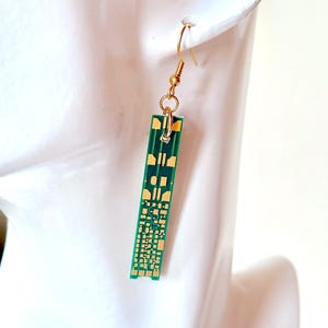 May include: A single gold-colored earring with a green circuit board design. The earring is rectangular with gold-colored accents and a gold-colored hook. The earring is hanging from a white surface.
