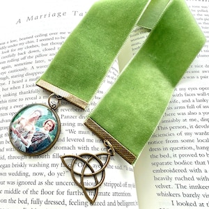 Op de afbeelding: Een lichtgroene fluwelen lintbladwijzer met twee bevestigde metalen bedels. De ene bedel is een ronde hanger met een gedrukte afbeelding van een koppel in historische kleding, de andere is een bronzen metalen triquetra-knoopsymbool.