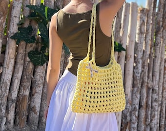 May include: A yellow crochet tote bag with a rectangular shape and open weave design. The bag has two handles and a small ribbon with text. The bag is held by a person wearing a green tank top and white skirt.
