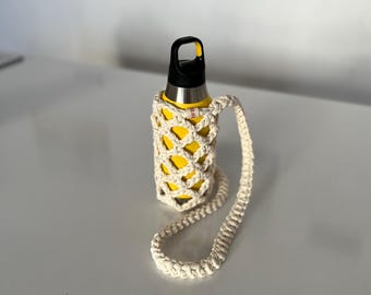 Puede incluir: Una botella de agua amarilla con tapa negra y un soporte de macramé color crema con una correa a juego. La botella es de acero inoxidable. El soporte es de algodón tejido.