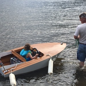 May include: A young person wearing a life vest is driving a small wooden boat with a white and gray hull. The boat has a small outboard motor and is being towed by an adult in the water.