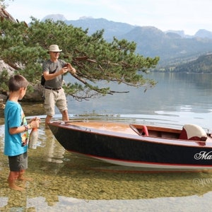 Puede incluir: Un pequeño bote de madera con un esquema de pintura blanco y negro está atracado en la orilla de un lago. El bote tiene un pequeño motor fuera de borda y el nombre "Matyi" está escrito en el lateral. Un hombre y un niño están de pie en la orilla cerca del bote.