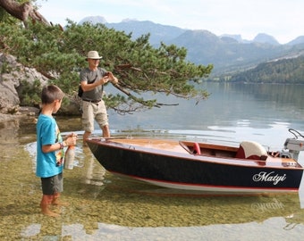 May include: A small wooden boat with a black and white paint scheme is docked at a lakeshore. The boat has a small outboard motor and the name "Matyi" is written on the side. A man and a boy are standing on the shore near the boat.
