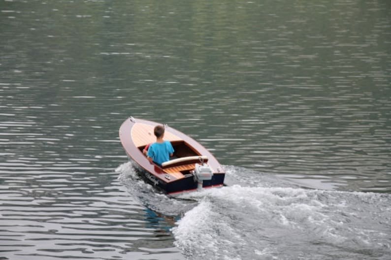 May include: A person in a blue shirt is driving a small wooden boat with a motor on a lake. The boat is creating a wake behind it.