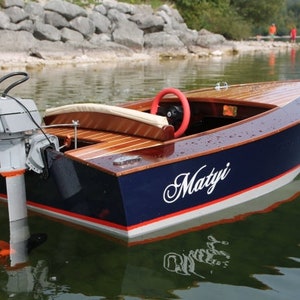 May include: A small wooden boat with a blue hull, white trim, and a red stripe. The boat has a wooden interior and a small outboard motor. The boat is named "Matyi" in white lettering on the side.