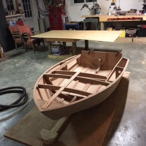 May include: A wooden boat hull under construction in a workshop. The boat is upside down and resting on a wooden stand. The boat is made of plywood and has a rounded shape.