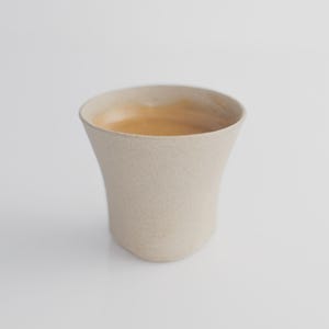Puede incluir: Una taza de café de cerámica beige con una superficie texturizada. La taza tiene un diseño curvo y acampanado y está llena de un líquido marrón claro, probablemente café o espresso. La taza está sobre un fondo blanco.
