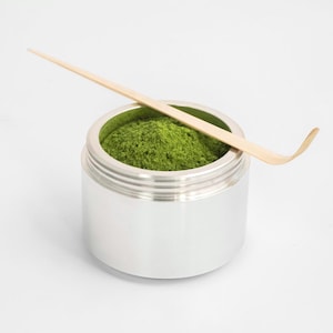 May include: A silver container filled with vibrant green matcha powder, topped with a light-colored bamboo whisk. The container has a ridged design and sits on a white surface. The whisk is angled across the top of the container.