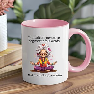 May include: White ceramic mug with a pink interior and handle. The mug features an illustration of an elderly woman in a yoga pose, with text that reads "The path of inner peace begins with four words. Not my fucking problem."